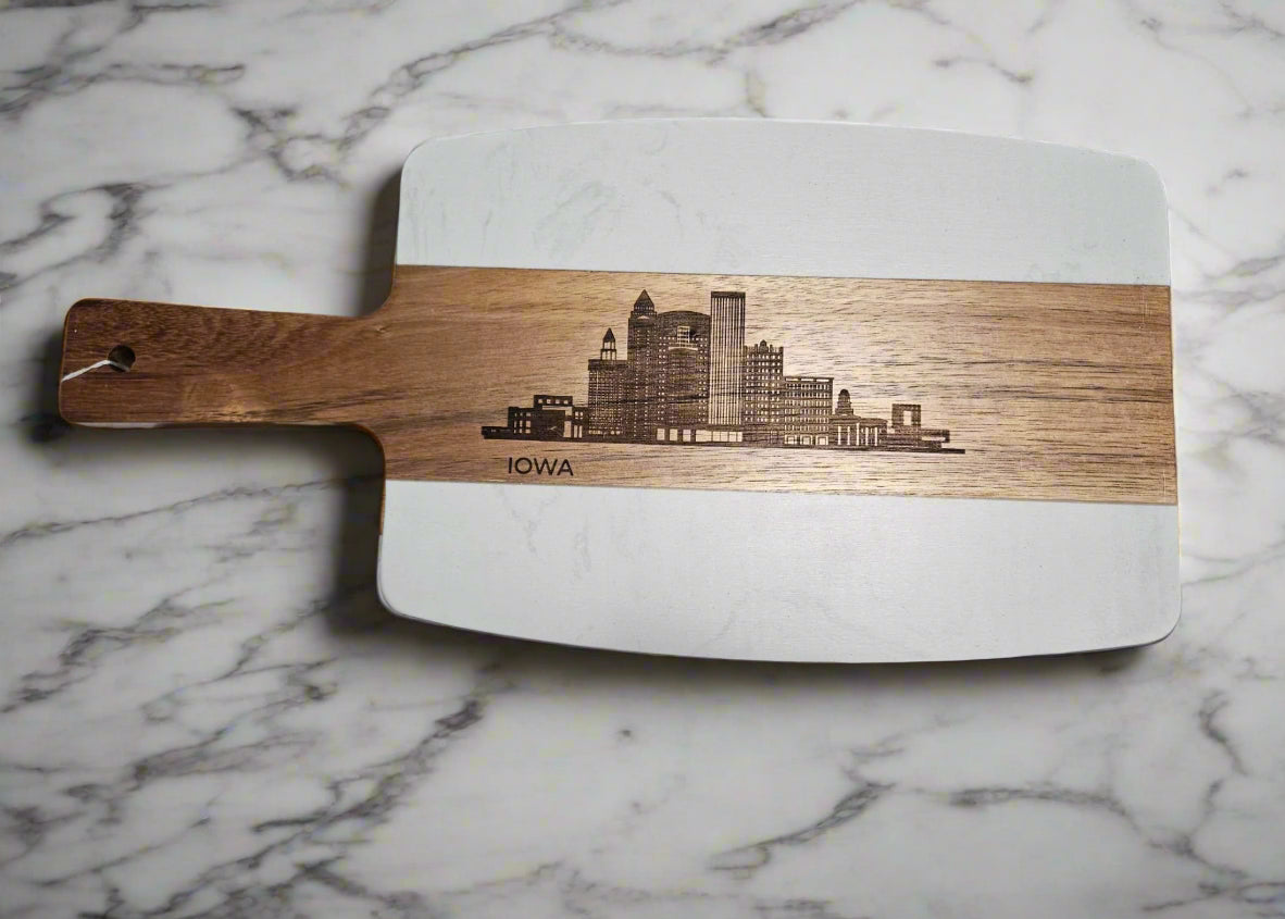 Wooden cutting board with laser-cut design of a city skyline and 'Iowa' text on a patterned fabric background.